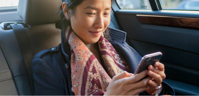 A young woman sits in the back seat of a car, smiling as she looks  down at her smartphone which she holds in both hands. She has long dark hair and is wearing a colorful patterned scarf.
